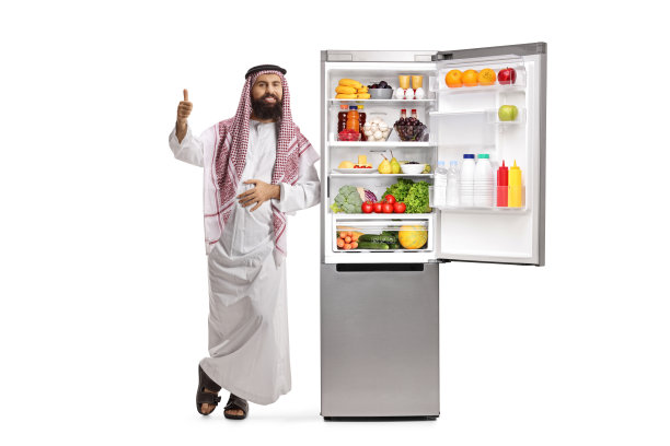 沙特阿拉伯男人的袋子靠在一个装满食物的打开的冰箱上，并竖起大拇指图片下载