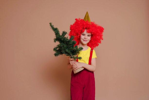 滑稽的小丑女孩戴着红色假发，举着一棵圣诞树，背景是香槟花。假期,马戏团的概念。图片下载
