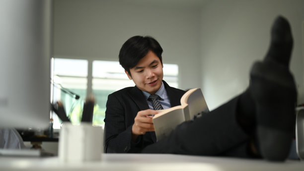 一位快乐成功的年轻经理正斜靠在椅子上，脚搁在桌子上看书。图片下载