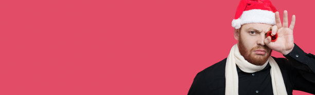 一名严肃的白种人蓄着胡子，戴着圣诞帽，用圣诞装饰球遮住眼睛，在一堵有自由空间的红墙上摆姿势图片下载