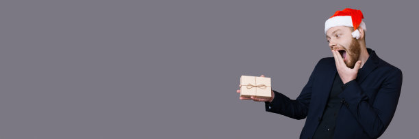 一名留着胡子的兴奋的白人男子正拿着一份礼物，捂着嘴在灰色的墙壁上做着幸福的手势图片下载