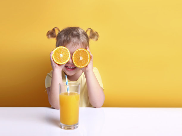 眼睛戴着橘子的小女孩坐在空的桌子旁，手里拿着一杯橙汁。健康维生素儿童营养。孩子与水果。图片下载