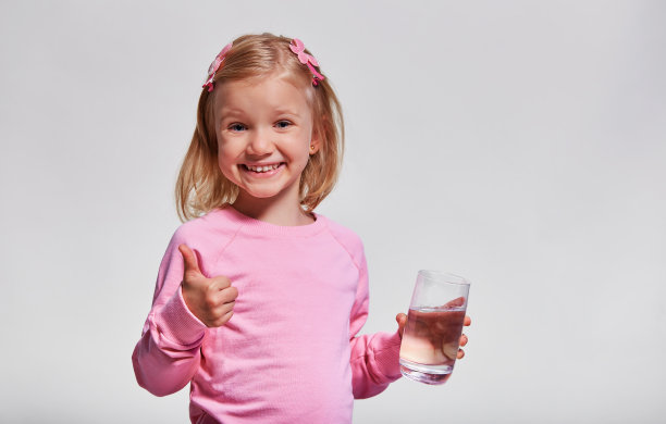 可爱的小女孩拿着一杯水。饮用水。肖像视图配置文件。孩子手里拿着玻璃，看着灰色背景的摄像机图片下载