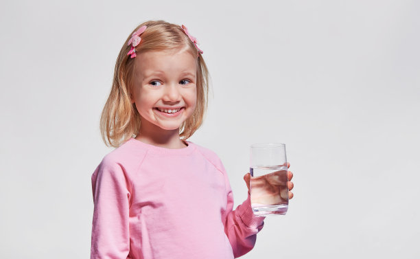 可爱的小女孩拿着一杯水。饮用水。肖像视图配置文件。孩子手里拿着玻璃，看着灰色背景的摄像机图片下载