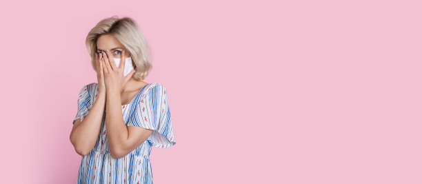 吓坏了的白种金发女人用医疗面具覆盖她的脸与手掌看在一个粉红色的工作室墙壁上的相机与自由空间图片下载