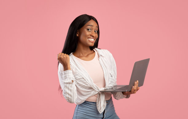 千禧一代非洲裔美国女士用笔记本电脑做YES手势，并在粉红色的工作室背景上微笑图片下载