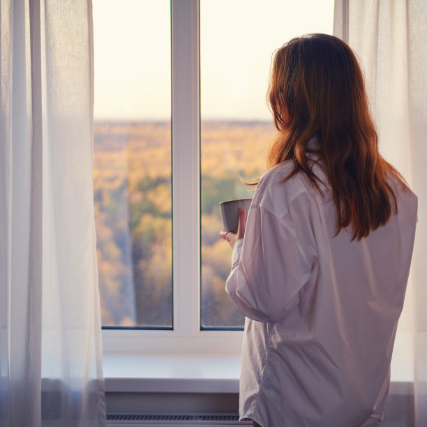一个女人站在窗前，俯瞰着秋天的森林。一个手里拿着杯子的年轻女子看着这对有黄色树木的夫妇图片下载
