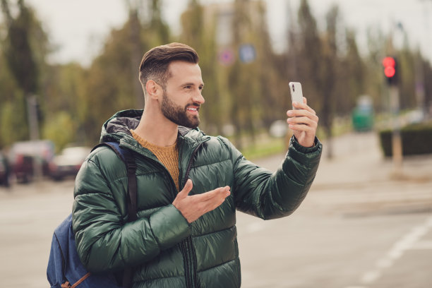 自信迷人的年轻人穿着绿色风衣书包在城市街道外传递现代设备的照片图片下载