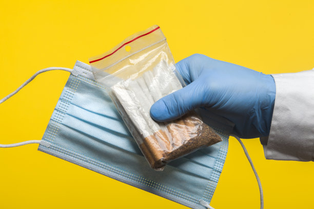 佩戴防护手套的医生手持一个密封的铝箔袋，里面装着香烟和防护口罩，以防止冠状病毒感染COVID-19，隔离在黄色背景上图片下载