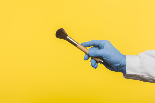 戴着蓝色防护手套的女孩拿着眉毛和化妆刷。孤立在黄色背景上图片下载