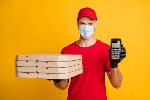 照片快递员拿着纸箱披萨盒终端穿红色t恤帽子手套孤立黄色背景图片下载