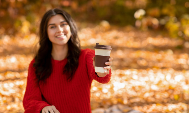 可爱的年轻女士在温暖的羊毛衫喝新鲜的热咖啡在秋天公园，选择性的焦点。本空间图片下载