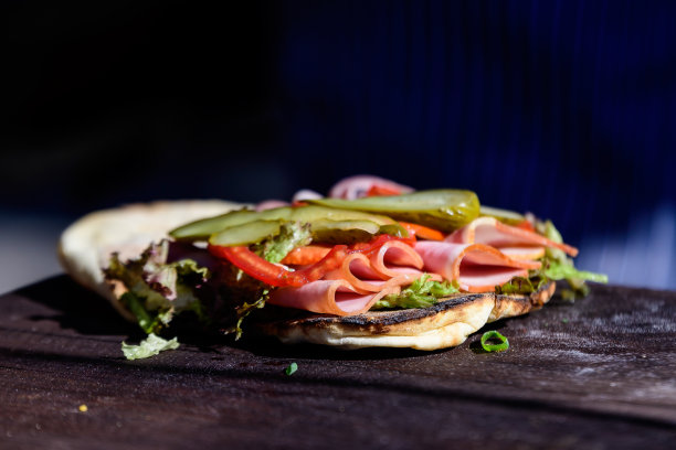 阳光直射下，一个新鲜健康的三明治或汉堡，夹着火腿、蔬菜沙拉和腌黄瓜，放在街边小吃市场的棕色木桌上，这是一张柔焦的侧视图照片图片下载