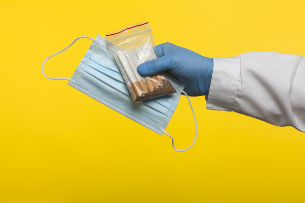 医生戴着防护手套，手持密封的铝箔袋，装有香烟和蓝色防护口罩，隔离在黄色背景上图片下载