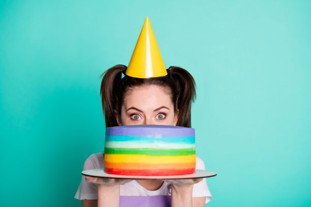 可爱害羞的女孩的照片，拿着条纹蛋糕隐藏脸看相机穿紫罗兰整体纸锥帽孤立的绿松石色背景图片下载