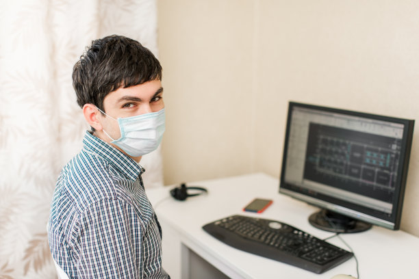 一个年轻人在他工作的地方用电脑在桌子旁工作。隔离期间在家工作戴着面具图片下载