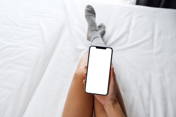 一名女子躺在舒适的白色床上，手里拿着白色手机，桌面屏幕是空白的图片下载
