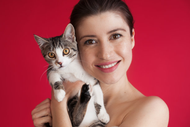 一名长着雀斑、裸露肩膀的白种女性在镜头前展示了一只微笑的猫，她拥抱着一只猫，这只猫挂在工作室的红色墙上图片下载