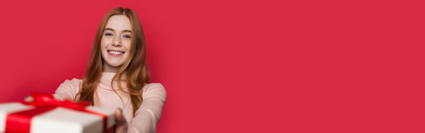 一个长着雀斑和红头发的可爱女人正对着镜头微笑，她正在接受一份放在工作室红色墙壁上的礼物，墙上有自由空间图片下载