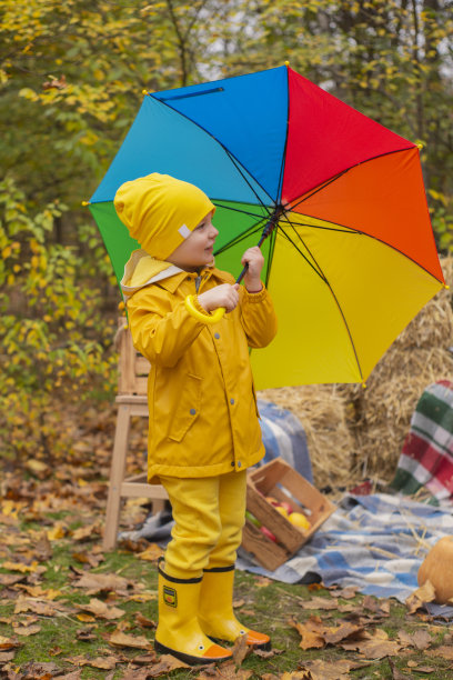 可爱漂亮的学龄前男孩，穿着橙色的裤子，雨衣，帽子，橡胶靴和彩虹色的雨伞附近的照片区秋季装饰品-南瓜，苹果，毯子，干草。舒适图片下载