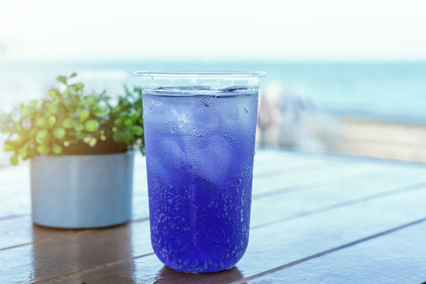 一杯混合着柠檬汁和紫色豌豆花的冰水和水滴放在前面模糊的蓝色海洋背景与复制空间的桌子上——假日和健康饮料的概念。图片下载