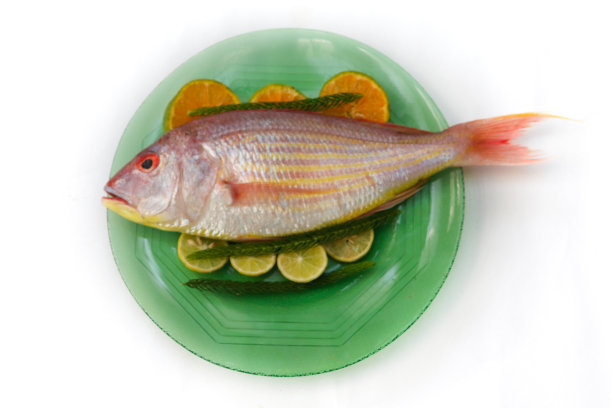 近距离观察新鲜的粉红色鲈鱼(线鳍鲷)装饰有柠檬片，橙色片和咖喱叶在玻璃盘子上，白色背景，选择性焦点。图片下载