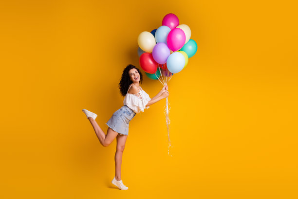 全尺寸的档案照片，乐观甜美的棕色卷曲的女孩抱着许多气球，穿白色上衣蓝色裙子运动鞋孤立在黄色的背景图片下载