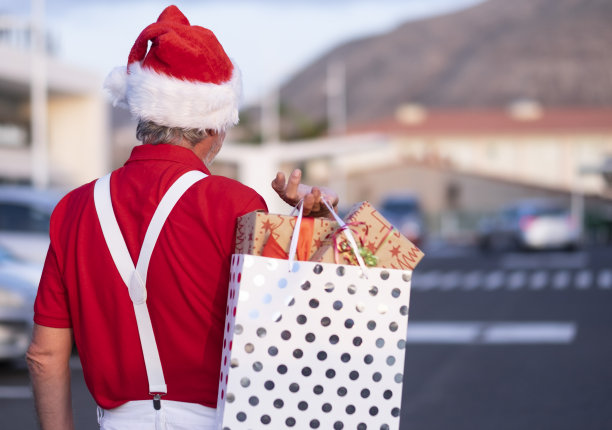 后视图，一个年迈的祖父购物与圣诞老人的帽子和吊带，抱着一个购物袋满礼物图片下载
