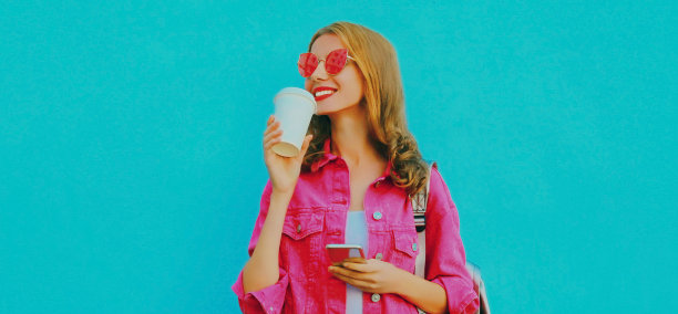 这是一幅时尚的年轻女子穿着粉色夹克，戴着墨镜，拿着智能手机喝咖啡的照片图片下载