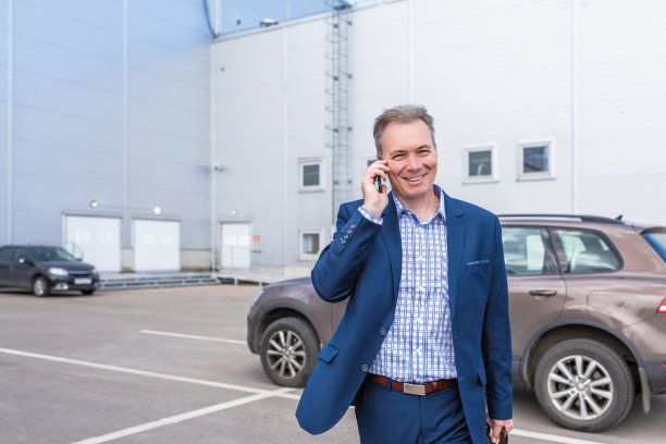 在一家大型工厂的私人停车场上，一位身穿蓝色西装的中年商人一边打电话一边笑着从他的车里走出来。图片下载