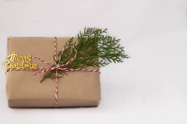礼盒用棕色牛皮纸包裹，用红白线系住，用天然松枝叶装饰。圣诞快乐，金色文本主题。白色背景与空白拷贝空间图片下载