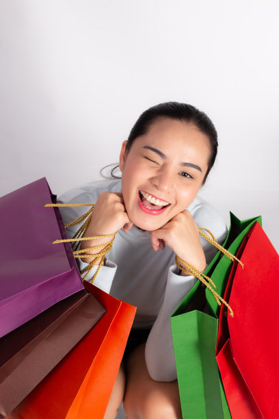 开朗美丽的亚洲女人拿着多种颜色的购物袋和显示ok标志在浅白色的背景。图片下载