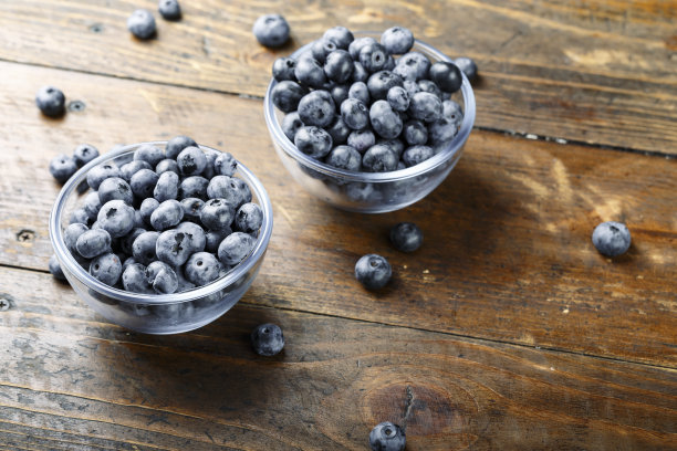 新鲜的蓝莓放在一个玻璃碗里，散落在一张木桌上。食物含有抗氧化剂。图片下载