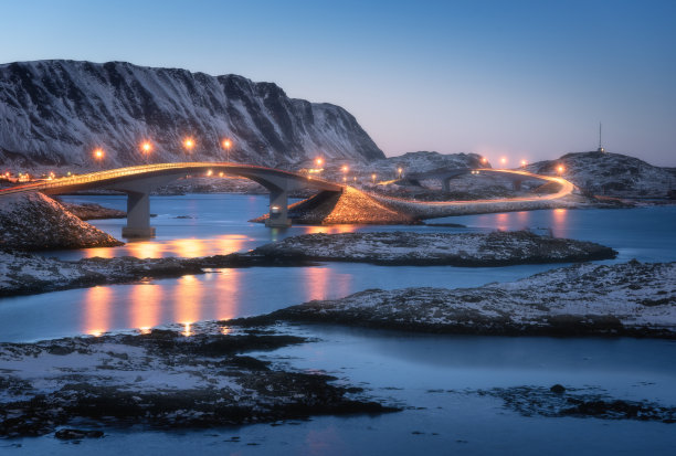 桥上灯火通明，雪山覆盖，村庄和蓝天映在水中。夜景有桥、雪石、城市灯光、大海。挪威罗浮敦群岛的冬天。路图片下载