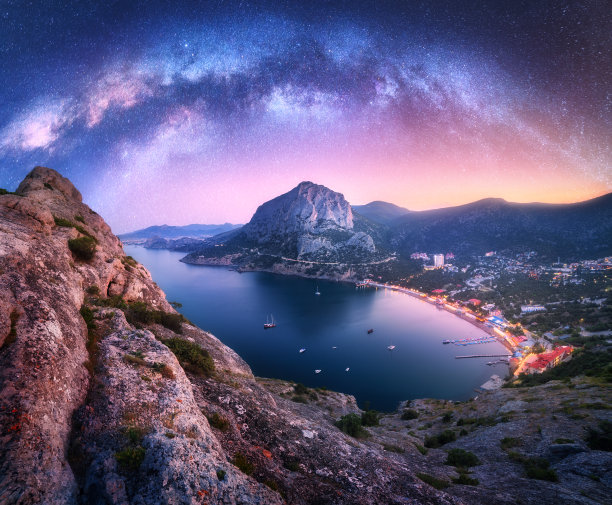 在夏天的夜晚，拱形的银河越过美丽的山脉和海岸。多彩的景观有岩石，明亮的星空，银河拱门，城市灯光，船只和游艇。星系。性质和空间图片下载