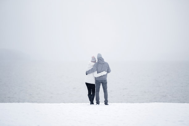 一对年轻的成年夫妇站在雪地上，在湖边互相拥抱，遥望远方。浪漫可爱的时刻。白色寒冷多雪的冬日。和平的气氛。约会的概念。后视图。图片下载