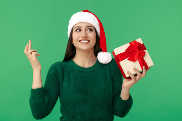 漂亮的年轻的黑发女人戴着圣诞老人的帽子在绿色的背景希望交叉手指。图片下载