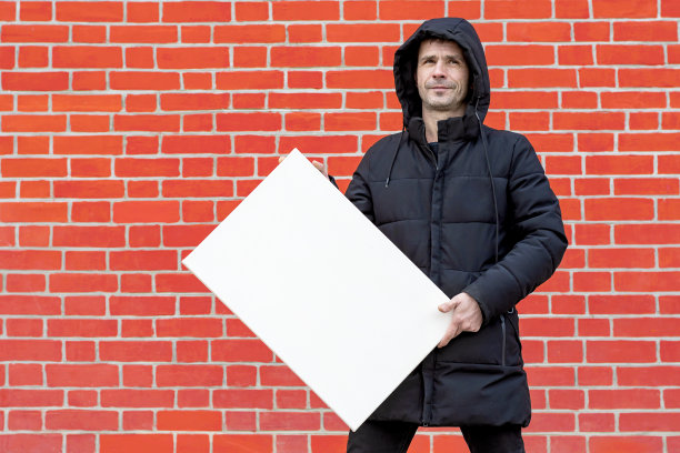 一个穿黑衣服的人靠在一堵红砖墙上，展示着一块空白的白画布图片下载