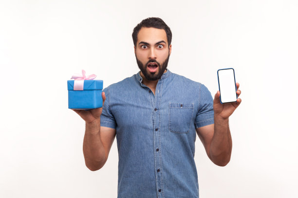 一名留着胡子的兴奋惊讶的男子拿着礼品盒和空屏幕的智能手机看着摄像头，大眼睛震惊了图片下载