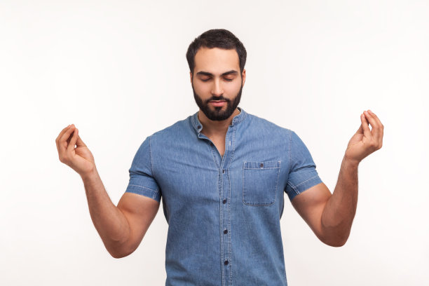 一名身穿蓝色衬衫、蓄着胡须的男子站在那里，紧闭双眼，紧紧地按在手上，正在练习瑜伽手印，以放松和集中注意力图片下载