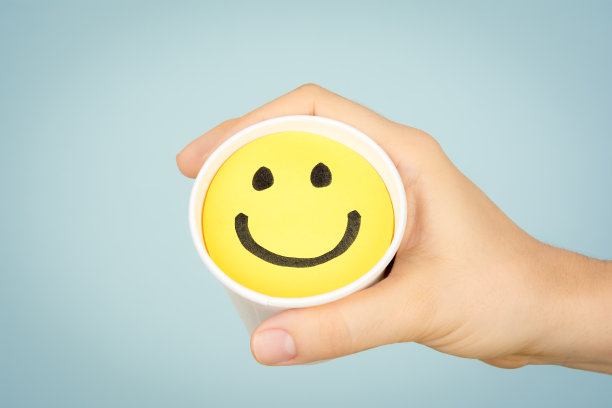 手握纸杯与一个快乐的微笑表情在黄色的纸和蓝色的背景。幸福的概念。图片下载