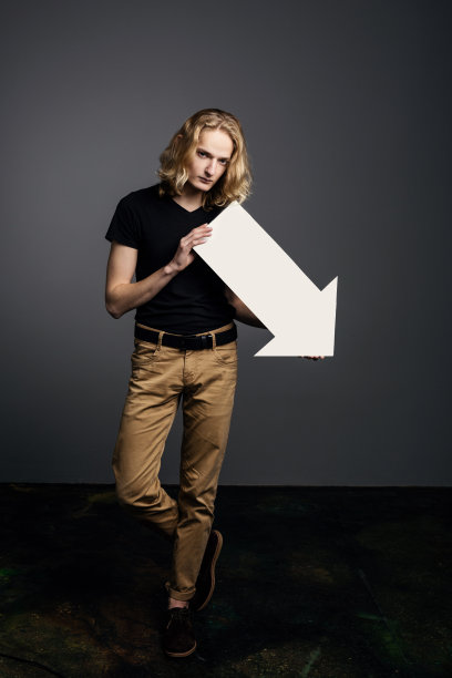 一个有着长长的金发的年轻帅气的男人，手里举着一个巨大的白色箭头，箭头指向下方，灰色背景上的一张沮丧的脸图片下载