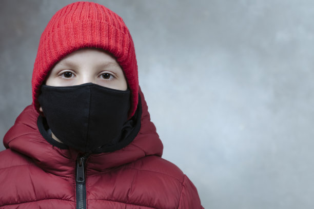 男孩戴着冬季防护冠状病毒的安全黑色口罩，红色夹克和红色帽子，背景受损。特写。Covid-19在全球爆发图片下载