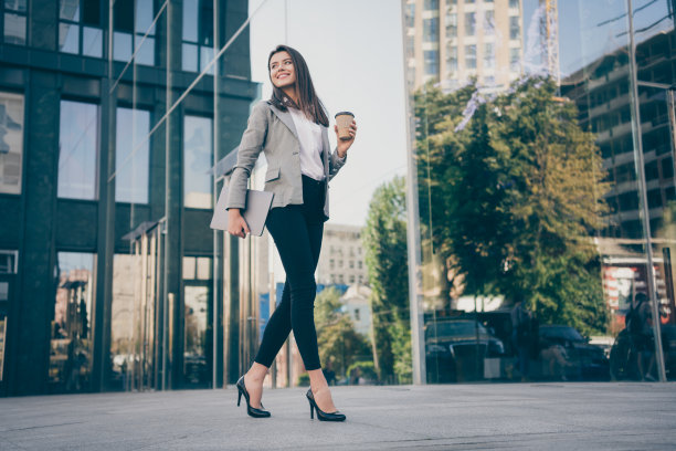全尺寸的照片，年轻的商务女士拿着笔记本电脑，咖啡杯，穿西装走在户外大楼图片下载
