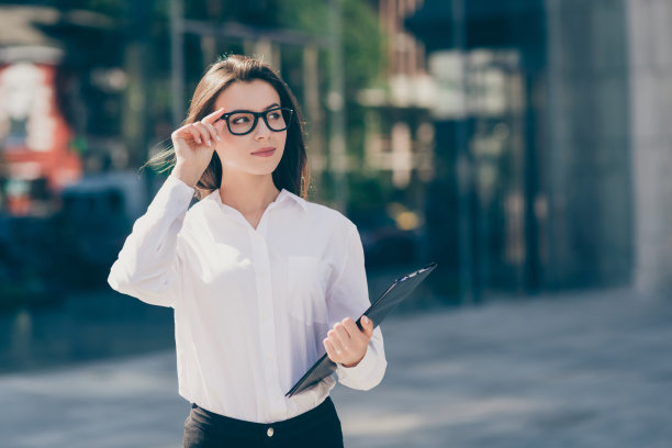 照片中的自信漂亮的年轻女子穿着白色正装衬衫，手戴眼镜，拿着写字板在户外图片下载