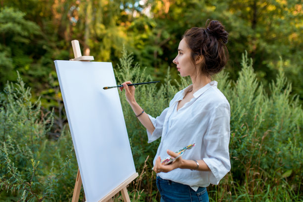 美丽的女子艺术家在夏天的公园，背景森林树木绿色的灌木丛，画的图片，在手调色板与油漆刷和画架。衣服白衬衫。白色的空白画布，开始绘画。图片下载