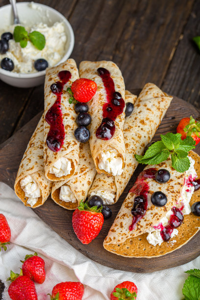 白软干酪裹在薄饼里，还有浆果、草莓、蓝莓、薄荷、果酱，上面和旁边的照片，放在一个木制砧板上，在一个黑暗的孤立的背景上图片下载