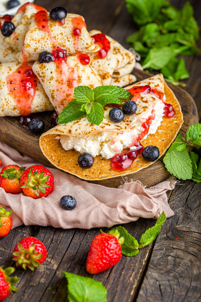 白软干酪裹在薄饼里，还有浆果、草莓、蓝莓、薄荷、果酱，上面和旁边的照片，放在一个木制砧板上，在一个黑暗的孤立的背景上图片下载