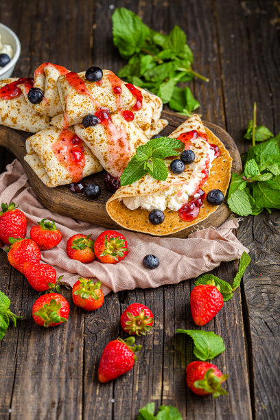 白软干酪裹在薄饼里，还有浆果、草莓、蓝莓、薄荷、果酱，上面和旁边的照片，放在一个木制砧板上，在一个黑暗的孤立的背景上图片下载