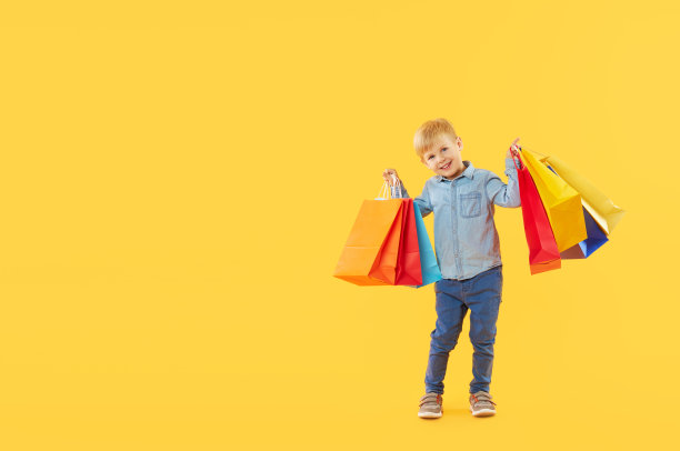 销售儿童产品。黄色背景上有趣的小男孩的肖像拿着购物袋，包装。看着摄像机。横幅。空间的文本。图片下载